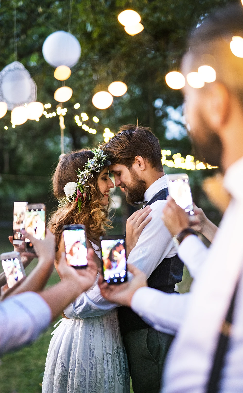 Newlywed couple embraces beneath glowing string lights in a garden as guests hold up smartphones, capturing their romantic wedding moment.