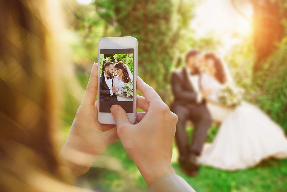 A person photographs a smiling bride and groom with a smartphone, framing the newlyweds in a romantic, sunlit garden wedding scene.