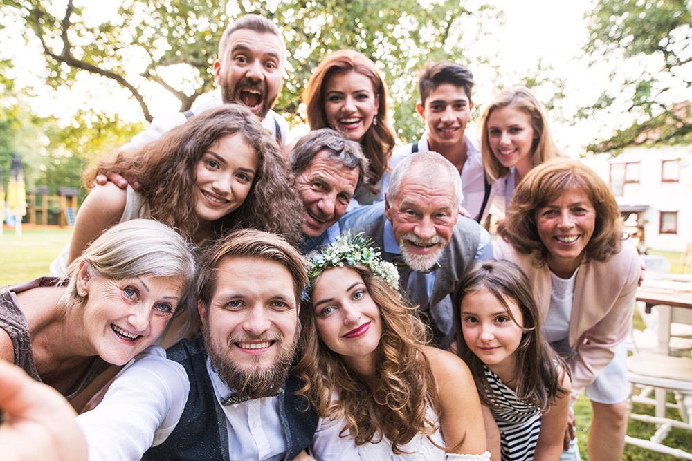 Diverse group of friends smile and laugh together in a sunny park, gathered closely for a joyful outdoor selfie with trees behind them.