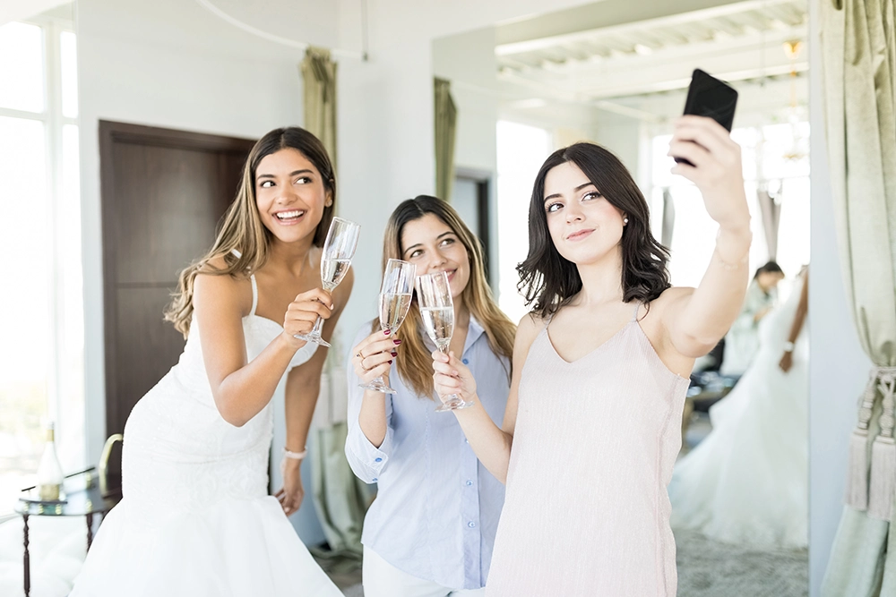 Drei Frauen heben bei einer ausgelassenen Hochzeitsfeier Champagnergläser und lächeln, während eine von ihnen ein Selfie macht; im Spiegel erscheint ein Brautkleid.