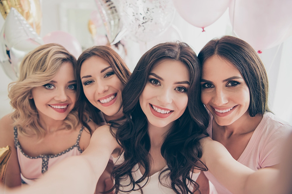 Four smiling women pose closely for a joyful selfie at a festive party, surrounded by pink and white balloons and soft celebratory décor.