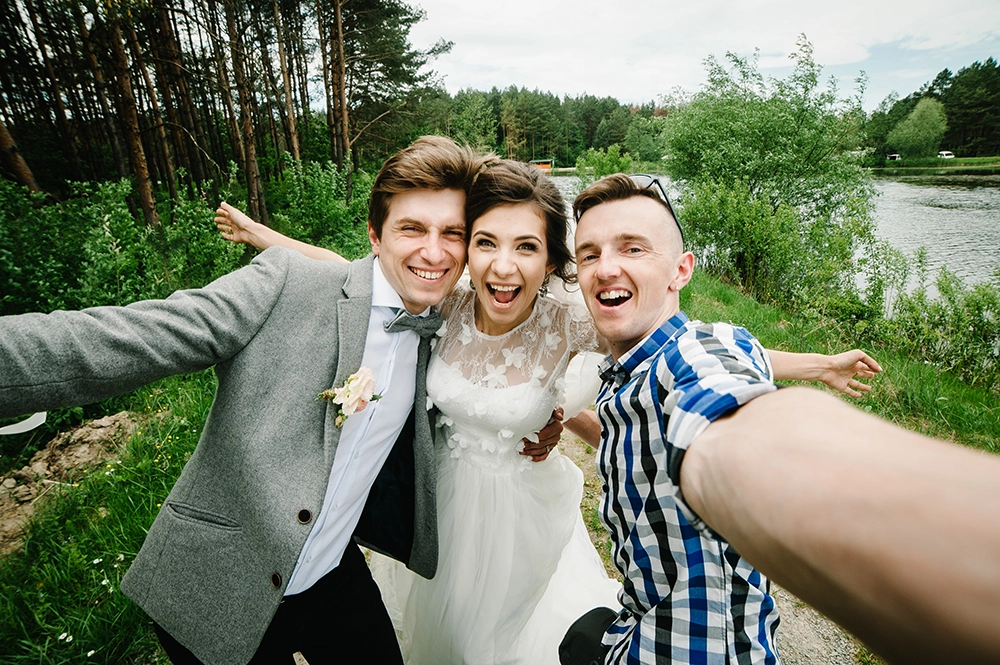 Un gioioso selfie di nozze che ritrae gli sposi e un amico in posa con le braccia aperte accanto a un lago immerso nel bosco, sorridenti e raggianti di gioia.