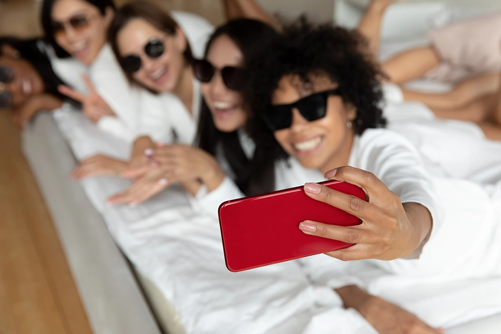 Friends in white pajamas and sunglasses smile for a joyful group selfie with a red smartphone while lounging together on a bed.