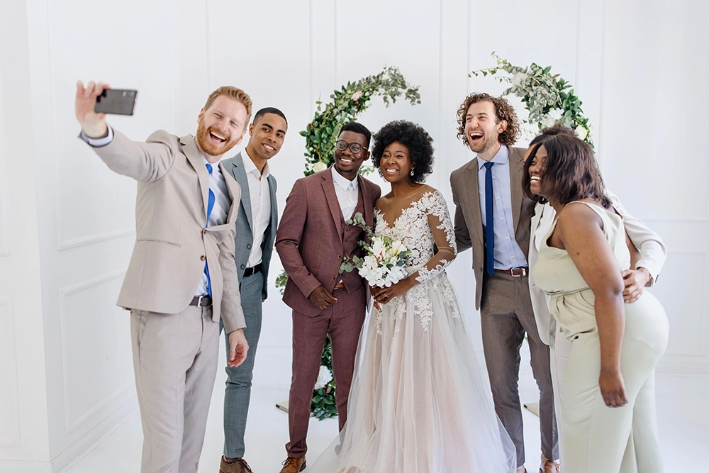 Diverse wedding party smiles for a joyful smartphone selfie, with the bride holding a bouquet beneath a greenery arch in a bright room.