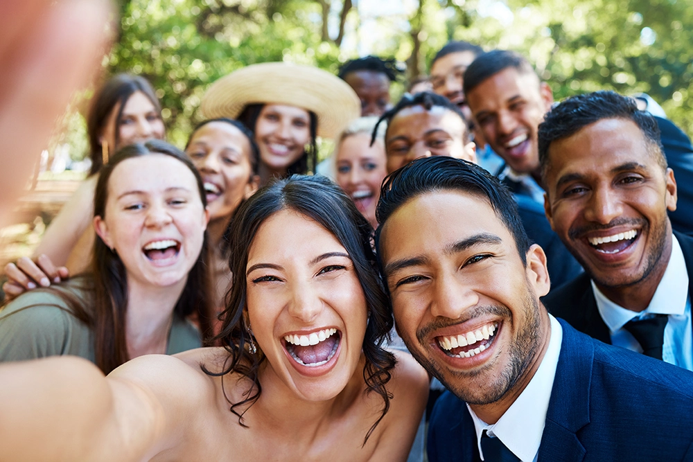 Joyful newlywed couple takes a selfie outdoors with a diverse group of friends, all smiling and laughing in bright natural light.