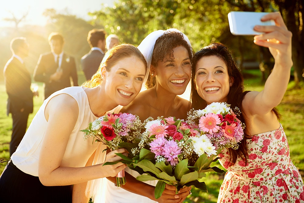 Three smiling women pose for a selfie in a sunlit garden, holding colorful flower bouquets; friends in the background create a joyful mood.