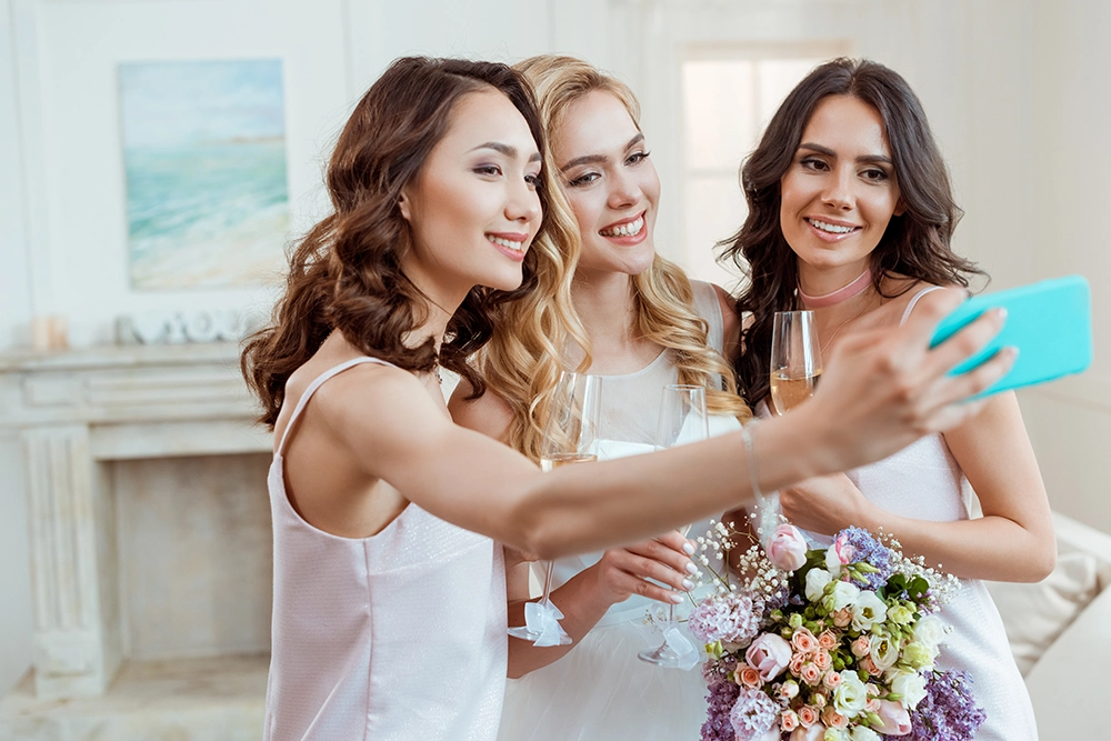Drei lächelnde Frauen in eleganten weißen Kleidern machen ein Selfie bei einer ausgelassenen Feier; sie halten Champagnergläser und einen bunten Blumenstrauß in den Händen.