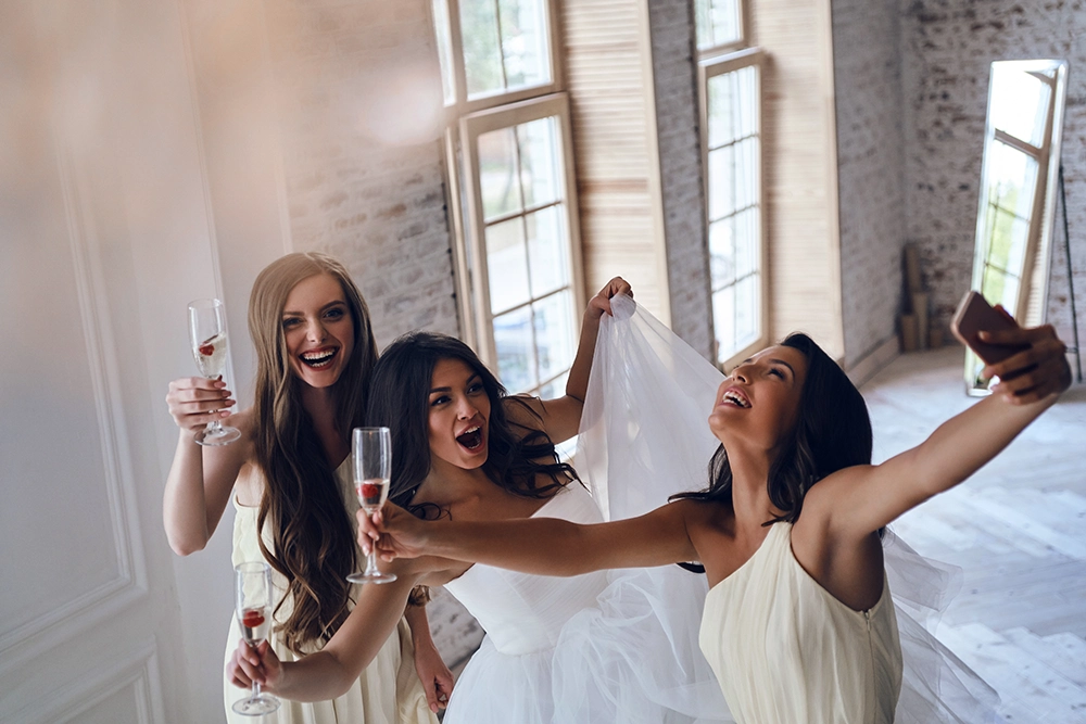 Three joyful women celebrate in a bright loft, raising champagne glasses and posing for a selfie as one wears a white bridal veil.