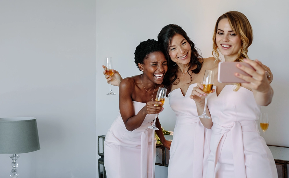 Three friends in pale pink dresses laugh and pose for a smartphone selfie while holding champagne glasses, creating a joyful celebration.