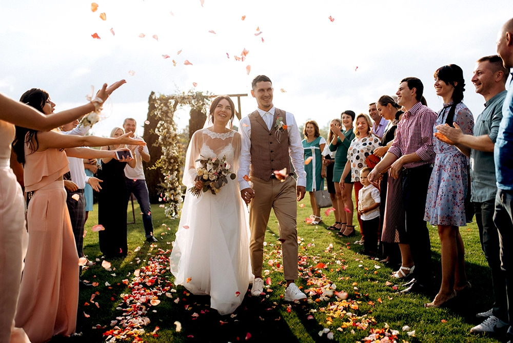 Bride and groom walk down a flower-strewn outdoor aisle as cheering guests toss petals, creating a joyful, celebratory wedding scene.