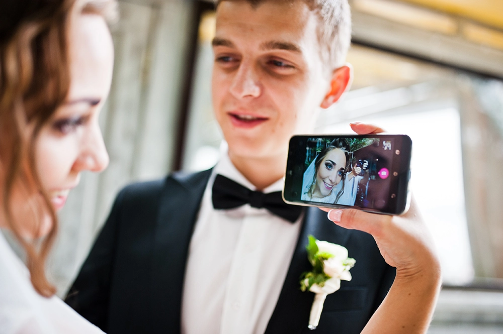 El novio, vestido con esmoquin, sostiene un teléfono inteligente en el que se ve a la novia sonriente, mientras ella mira hacia abajo a su lado, creando un tierno momento nupcial.