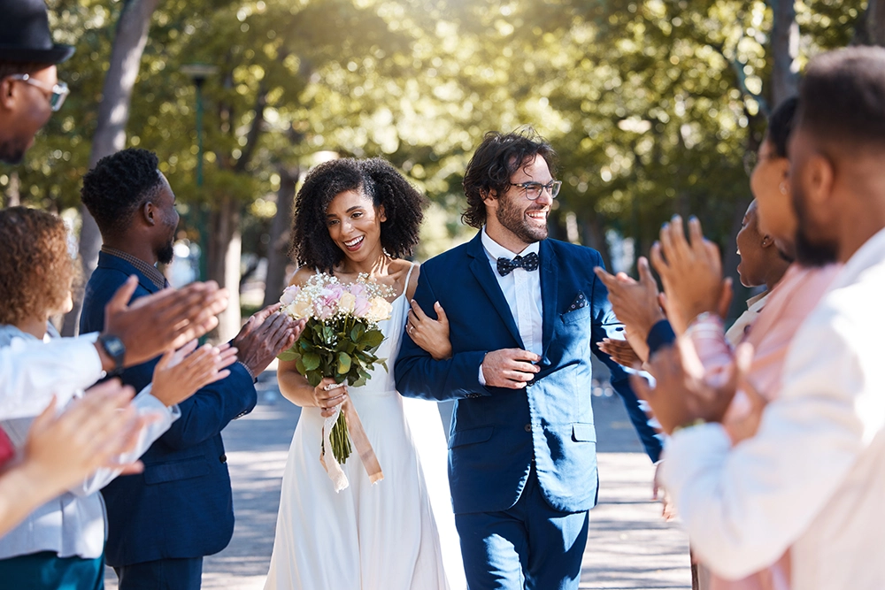 Una coppia di novelli sposi, sorridenti, passeggia in un parco illuminato dal sole tra gli applausi degli invitati; la sposa tiene in mano un bouquet e lo sposo indossa un abito blu.