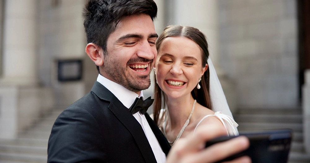 Newlywed bride and groom smile while taking a selfie outdoors, celebrating their wedding day with joy and affection.