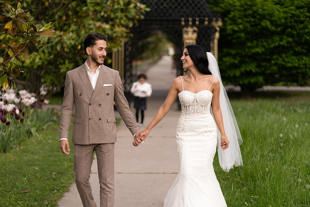 Gli sposi novelli sorridono mentre camminano mano nella mano lungo un sentiero in un giardino, circondati dal verde, dai fiori e da una soffusa luce romantica.
