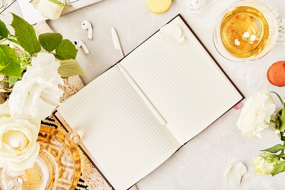 Un cuaderno abierto con papel cuadriculado en blanco, rodeado de flores blancas, hojas verdes, tazas de té, bastoncillos de algodón y detalles en tonos pastel, en una escena tranquila y elegante.