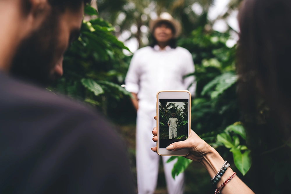A person photographs a smiling woman in white standing among lush tropical plants, creating a warm, joyful outdoor portrait scene.