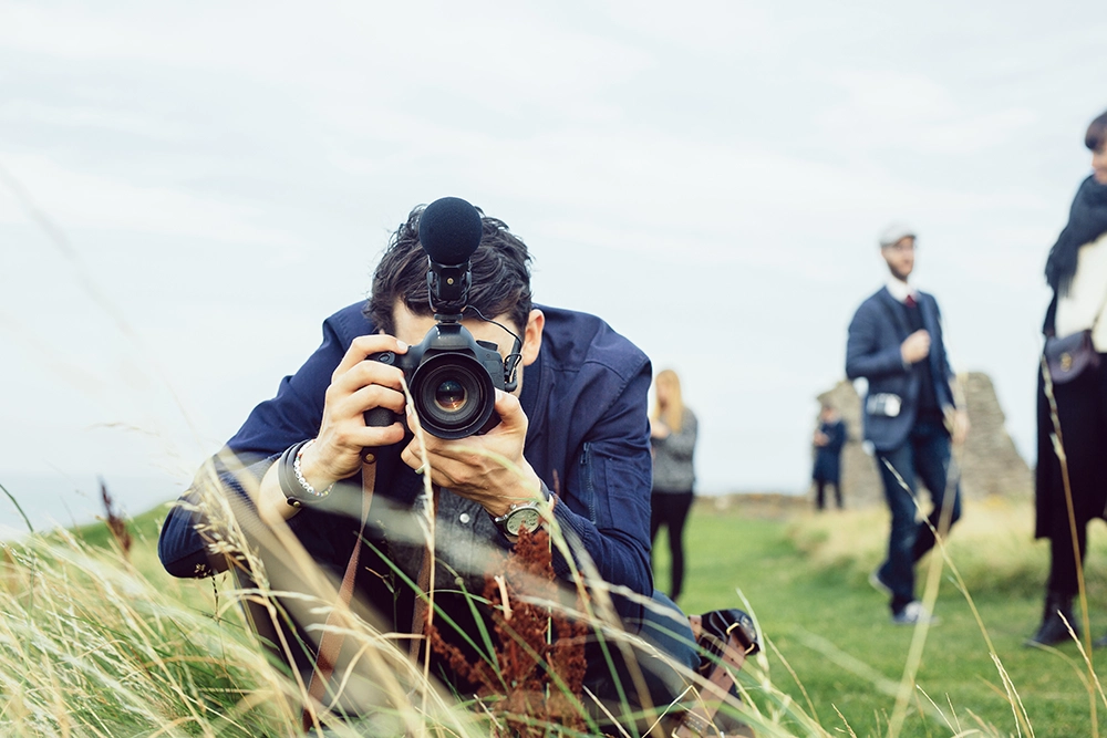 Photographer crouches in tall grass, focusing a DSLR camera, while formally dressed people stand blurred in the sunny background.