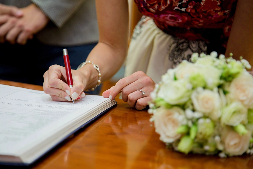 Bride signs wedding documents at a wooden table beside a bouquet of white and green flowers, capturing an intimate, joyful ceremony.