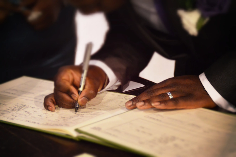 Two people in formal black attire sign a large guestbook, one guiding a pen across handwritten pages in a solemn, ceremonial scene.