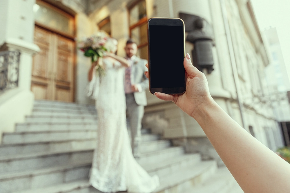 A hand holds a blank smartphone toward a softly blurred bride and groom on church steps, evoking wedding photography and celebration.