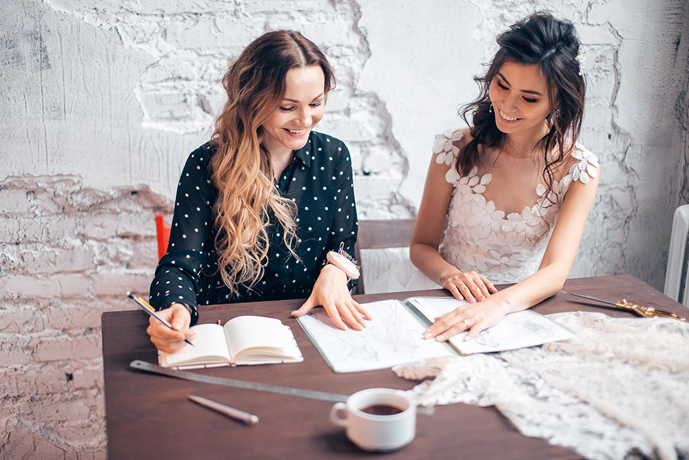Smiling bride and woman in a polka-dot blouse review wedding plans together at a rustic table, signing papers beside coffee and lace.