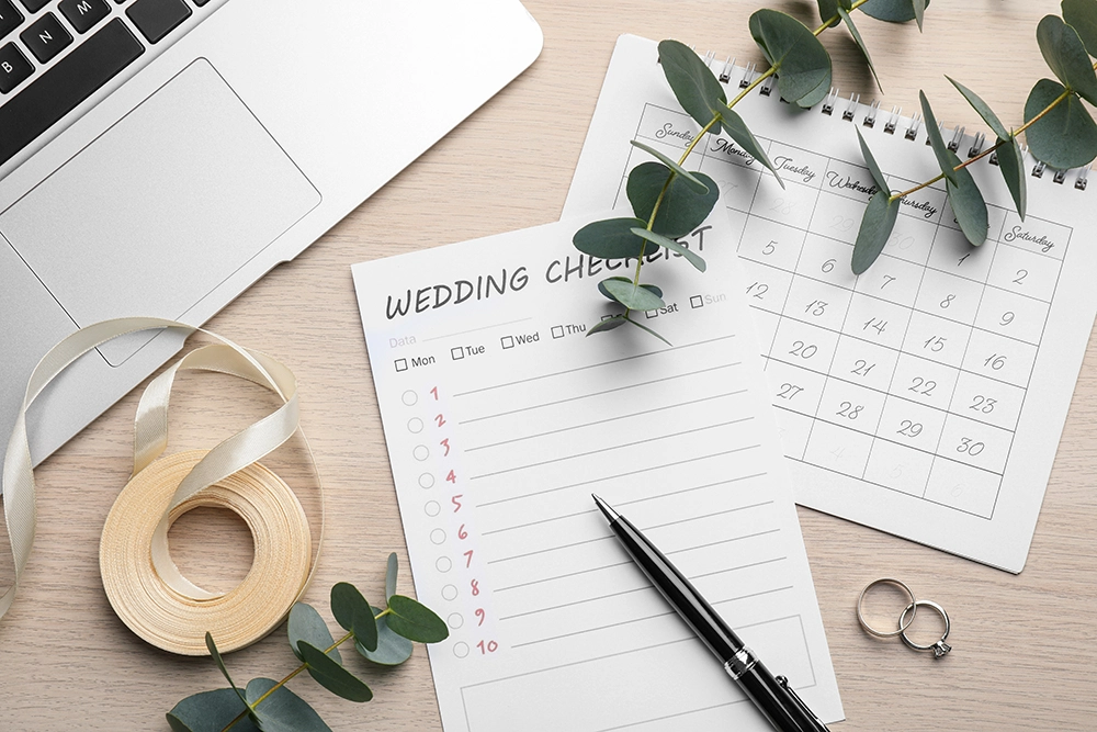 Wedding checklist, calendar, laptop, eucalyptus branches, ribbon, and wedding rings arranged on a light wood desk, conveying calm preparation.