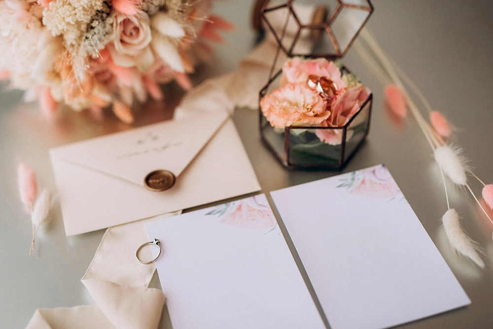 Romantic wedding stationery with blank floral cards, a sealed envelope, wedding rings, dried flowers, and geometric glass vases in soft pastel tones.