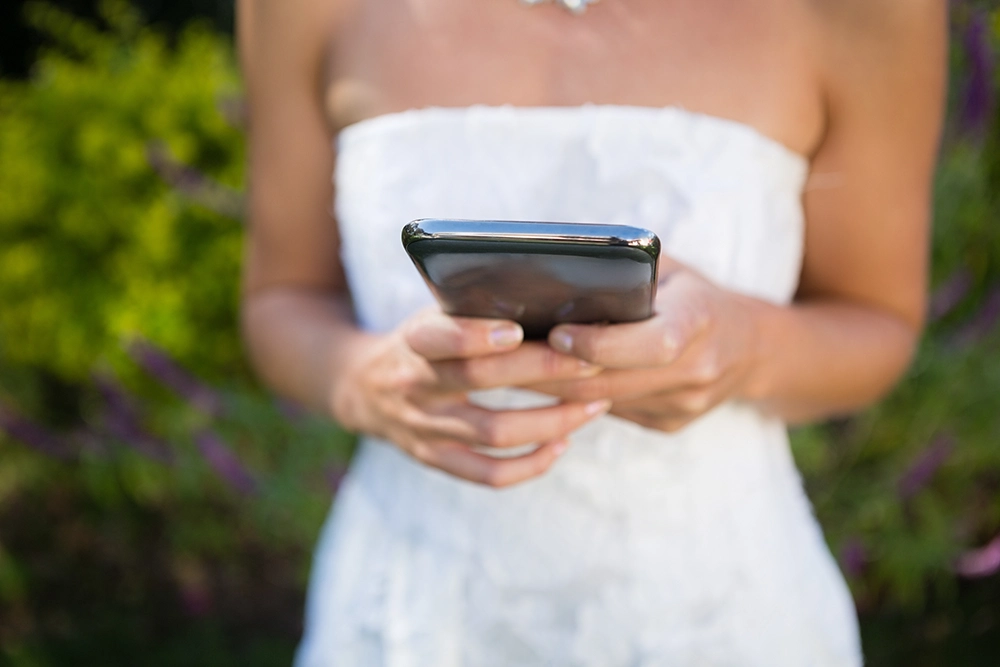 Woman in a white wedding dress holds a smartphone with both hands outdoors, suggesting wedding planning, communication, or a digital moment.