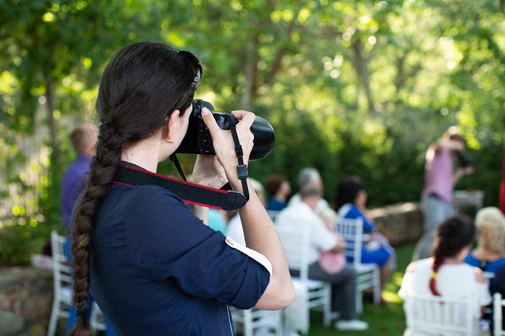 Photographer with a long braid aims a DSLR camera at an outdoor gathering, where seated people socialize among sunlit green trees.