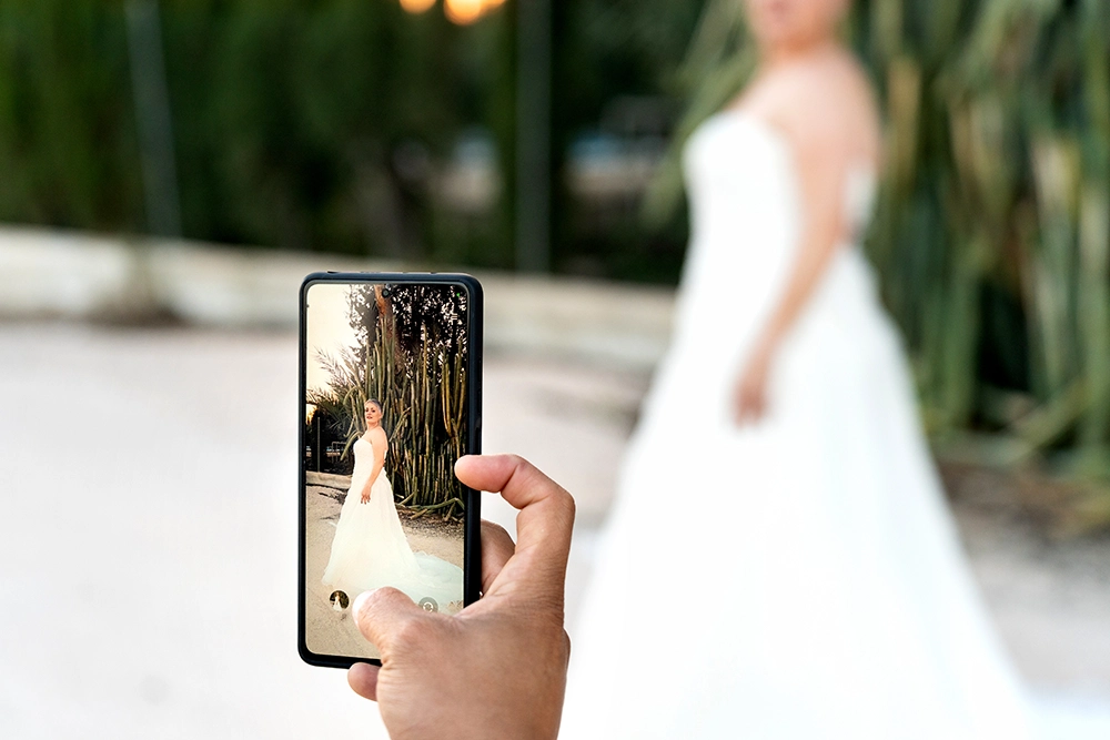 Una mano sostiene un teléfono inteligente que muestra a una elegante novia con un vestido blanco, mientras que la novia real aparece borrosa en un exuberante jardín, creando una atmósfera onírica.