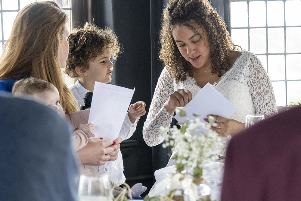 At a joyful wedding reception, a woman in a white lace top reviews papers with a curly-haired child as guests gather around a flowered table.