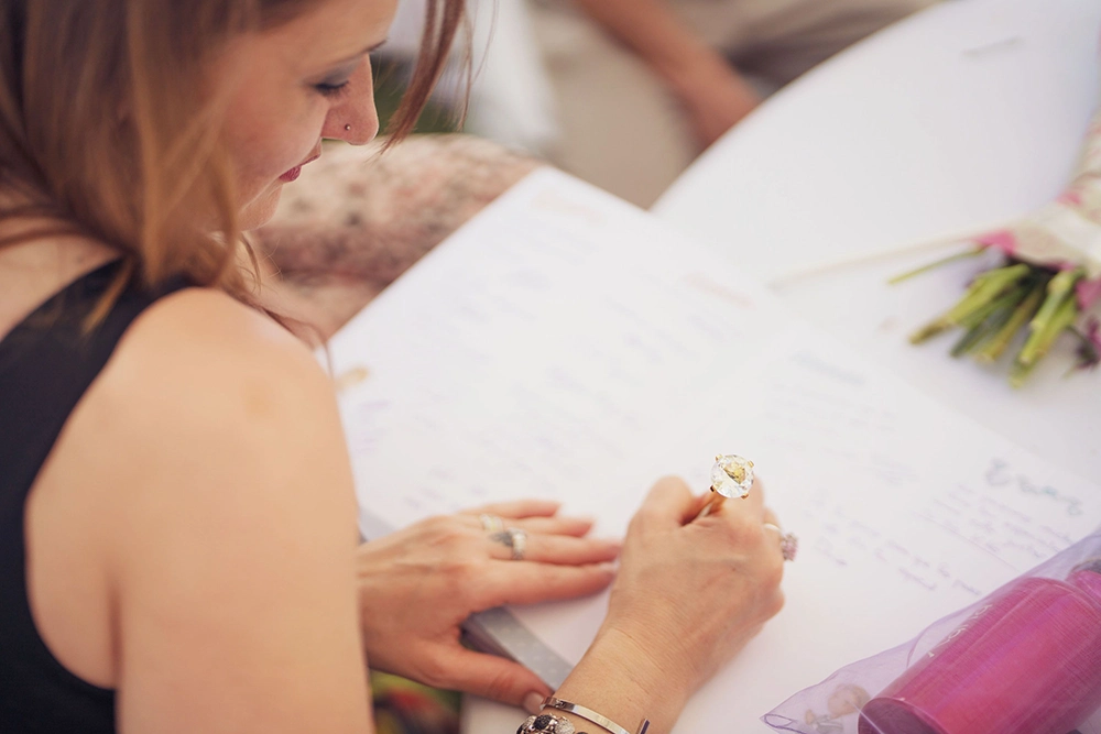 Woman in a black dress writes a heartfelt message in a wedding guestbook, with flowers nearby, creating a warm, intimate celebration mood.