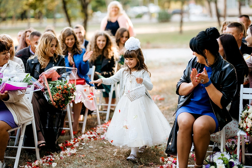 Young flower girl in a white dress walks down an outdoor wedding aisle as guests applaud, surrounded by rose petals and bouquets.